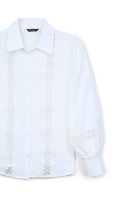 Camicia bianca con bottoni, decorata con pizzo lungo la parte anteriore e i polsini. Presenta un colletto classico e maniche leggermente voluminose.