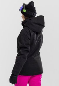 Personne portant une veste de ski noire, un bonnet noir avec un logo vert, des lunettes de ski bleues, des gants noirs et un pantalon de neige rose vif, vue de dos.