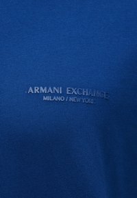 Μπλε ύφασμα με ανάγλυφο κείμενο που αναγράφει "ARMANI EXCHANGE MILANO / NEW YORK" στο κέντρο της επιφάνειας.