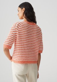Pull en tricot rayé en corail et blanc. Manches courtes, col rond, tissage texturé. Coupe courte avec ourlet et poignets côtelés.