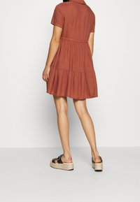 Robe à manches courtes de couleur rouille, en tissu doux, avec une jupe à étages, présentant un col et une taille ajustée. Associée à des sandales à plateforme noires.