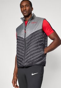 Muškarac nosi bezrukavu siva i crna podstavljena prsluk preko crvene majice kratkih rukava i crnih Nike hlača s bijelim logom, stojeći s rukom u džepu.