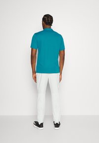 Türkises Poloshirt mit kurzen Ärmeln, weicher Stoff, Rückenansicht; kombiniert mit hellgrauen Hosen und schwarzen Sneakers. Einfaches Design und lässige Passform.