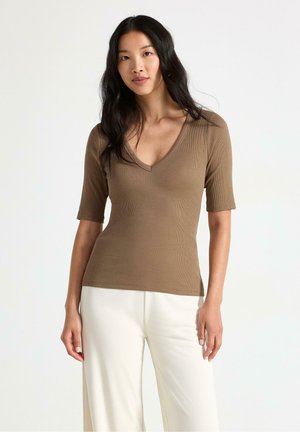Donna con lunghi capelli neri che indossa una maglietta marrone con scollo a V e maniche corte e pantaloni bianchi a gamba larga, in piedi davanti a uno sfondo semplice.