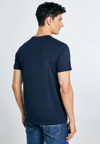 Man die met de rug naar de camera staat, gekleed in een effen marineblauw T-shirt met korte mouwen en een blauwe spijkerbroek, tegen een lichtgrijze achtergrond.