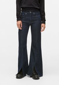 Jeans flare de denim escuro com fendas laterais, apresentando uma cintura alta, design de cinco bolsos e costura em contraste. Combinados com calçado preto.