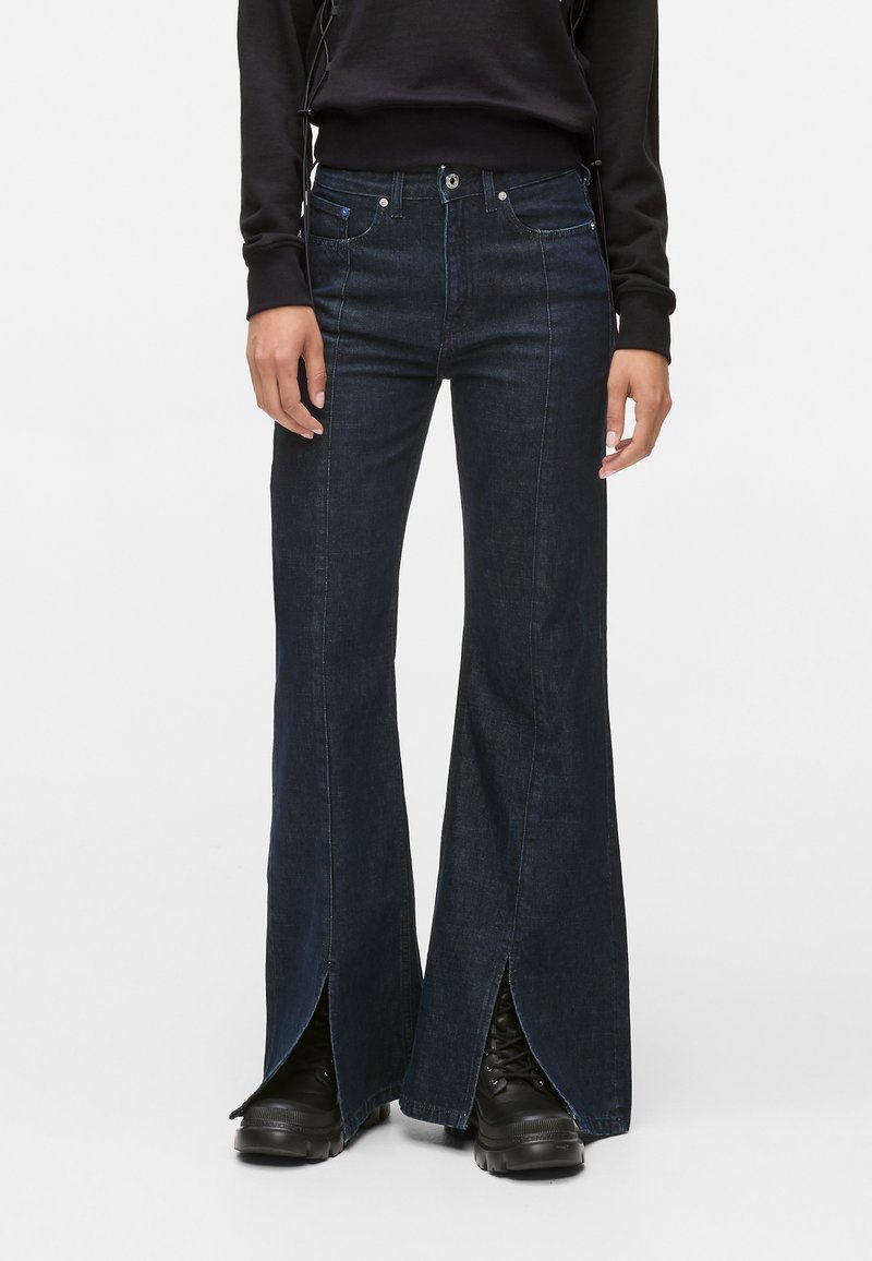 Jeans flare de denim escuro com fendas laterais, apresentando uma cintura alta, design de cinco bolsos e costura em contraste. Combinados com calçado preto.