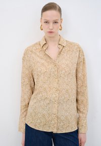 Beige košile na knoflíky s listovým vzorem, mírným leskem a dlouhými rukávy. Spárováno s tmavě modrými džíny, umístěné na bílém pozadí.