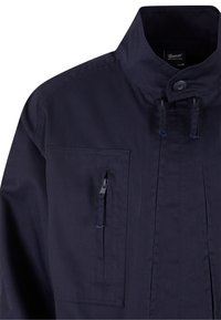 Veste bleu marine foncé avec col montant, fermeture par boutons, deux poches zippées verticales sur la poitrine et détails de couture visibles.