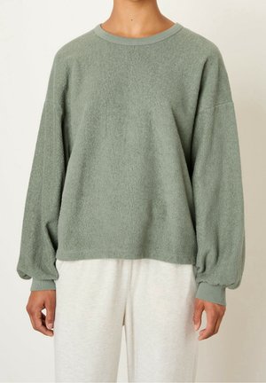 Personne portant un pull ample à manches longues vert clair duveteux et un pantalon blanc cassé, debout devant un fond clair uni.