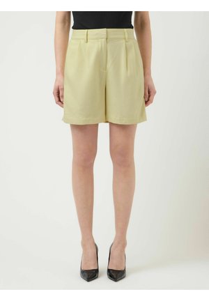 Vrouw draagt lichtgele, hooggetailleerde, op maat gemaakte shorts met plooien, een zwarte aansluitende top, en zwarte hoge hakken met puntige neus, staand tegen een witte achtergrond.