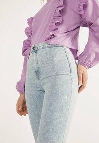 Lilakkinen paita, jossa on rypytys yksityiskohtia edessä ja hihoissa, yhdistettynä vaaleansinisiin korkean vyötärön farkkuihin, joissa on sileä pinta ja etunappi.