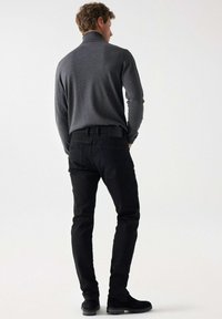 Siva majica s dugim rukavima i visokim ovratnikom uparena s crnim uskim trapericama i crnim cipelama, naglašavajući glatku teksturu i kroj prilagođen figuri.