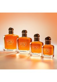 Vier bernsteinfarbene Parfümflaschen mit der Aufschrift Stronger With You Intensely von Emporio Armani, in einer Reihe auf einer reflektierenden Oberfläche angeordnet.