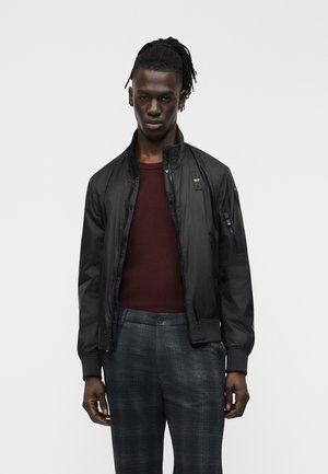 Jeune homme aux dreadlocks portant une veste noire zippée, un pull rouge foncé et un pantalon à carreaux gris, debout devant un fond clair uni.