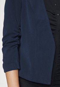 Marinblå jacka med mjuk textur, med trekvartslånga ärmar och en kortad design, lager-på-lager över en svart ribbad tröja med knappar.