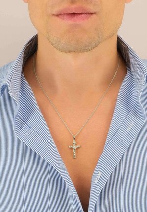 Pendente a forma di croce d'argento appeso a una catena delicata, indossato sopra una camicia a righe blu e bianche con colletto.