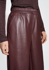 Pantaloni in pelle borgogna con vita elastica, tasche laterali e una superficie liscia, parzialmente visibili nella parte inferiore del corpo.