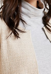 Béžová strukturovaná bunda s jemným povrchem, otevřený přední díl, nošená přes světle šedý rolák. Jsou viditelné přirozené kudrlinky vlasů.