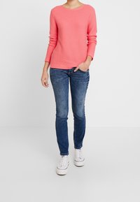 Koralowy sweter z długim rękawem w prążki, zestawiony z niebieskimi, przetartymi rurkami i białymi trampkami, stojący na jednolitym tle.