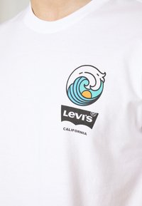 T-shirt en coton blanc avec un graphique de vague coloré en bleu et orange au-dessus d'un logo Levi's noir avec le texte "CALIFORNIA".