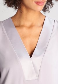 Blusa grigia chiara con scollatura a V profonda, realizzata in tessuto simile al satin. Presenta un design minimalista con leggera lucentezza e senza motivi visibili.