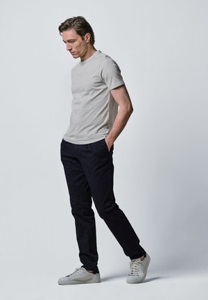 Mężczyzna w szarej koszulce, czarnych spodniach i białych sportowych butach stoi z rękami w kieszeniach, patrząc w dół na jednolitym jasnym tle.