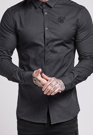 Mężczyzna w dopasowanej czarnej koszuli z długim rękawem, zapinanej na guziki, z haftowanym logo na piersi, splecione ręce ukazujące szczegółowe tatuaże.