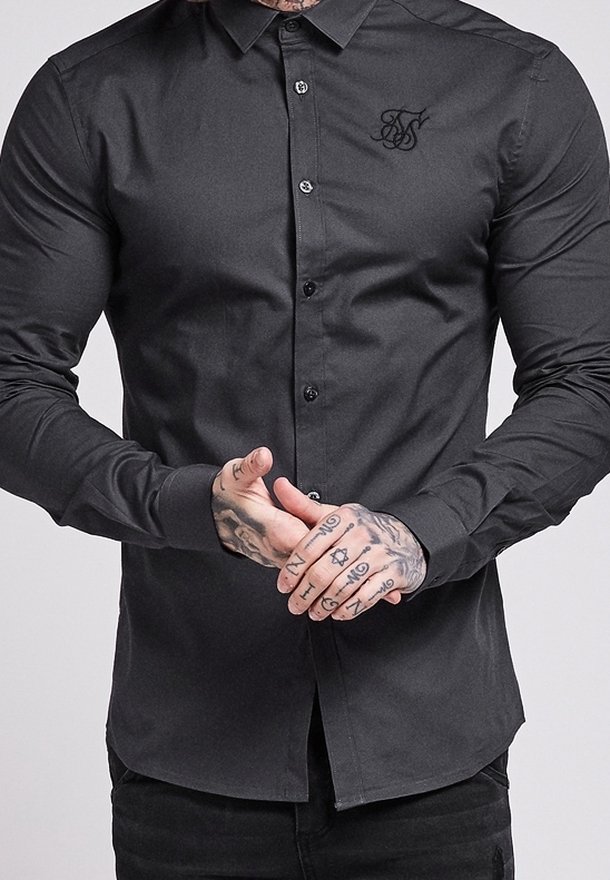 Homme portant une chemise noire ajustée à manches longues boutonnée avec un logo brodé sur la poitrine, mains jointes montrant des tatouages détaillés.