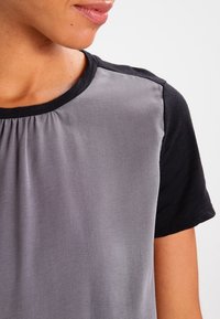 Kortärmad skjorta med en svart bomullstop och ett grått, silkesliknande tyg under. Halsringningen är rund med subtila veck vid sömmen.