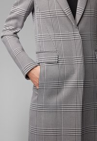 Abrigo a cuadros en gris y negro con un diseño ajustado, mangas largas y bolsillos frontales. La tela tiene una textura suave y un ajuste estructurado.