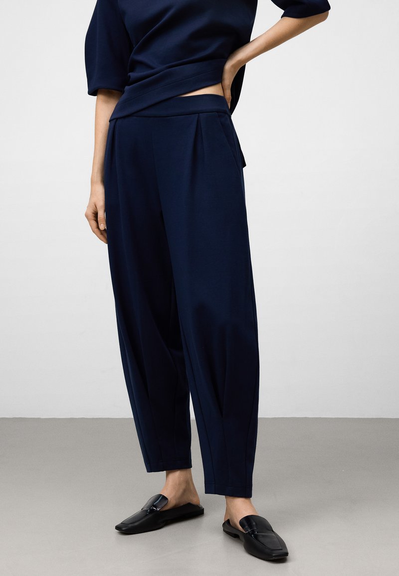 Vrouw in marineblauwe cropped wijde broek en bijpassende top, staande met één hand op de heup en zwarte leren instappers.