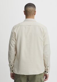Camisa de manga larga en tejido beige claro, con la espalda recta, puños abotonados y una textura suave con un acabado sutil.