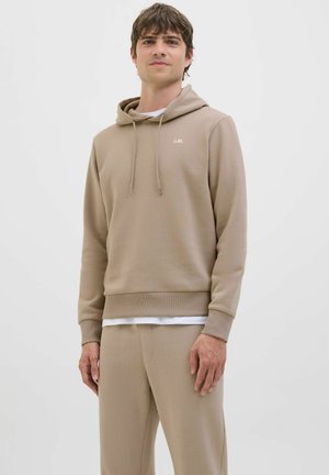 Joven con sudadera con capucha beige y pantalones deportivos a juego, de pie contra un fondo claro y liso, mirando al frente con expresión neutral.