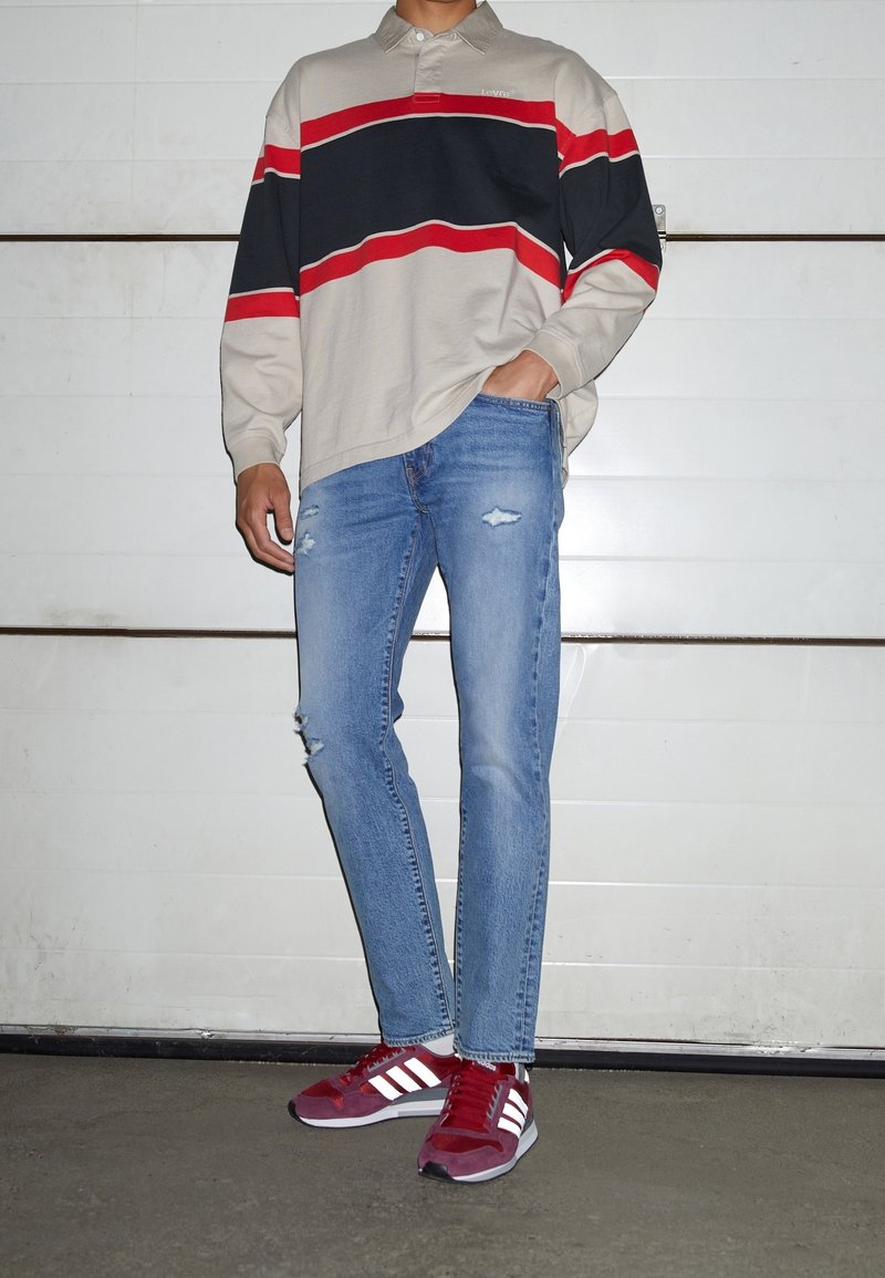 Personne portant un t-shirt à rayures beige, noir et rouge à manches longues, un jean bleu déchiré et des baskets rouges Adidas, debout contre un mur blanc.