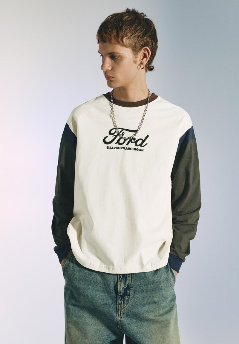 Jeune personne aux cheveux courts portant un shirt à manches longues blanc et foncé avec le texte « Ford Dearborn, Michigan » et un jean en denim, les mains dans les poches.