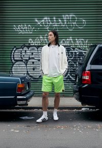 Biela chlpatá bunda s čiernymi pruhmi, biele tričko, neonovo zelené šortky s dizajnom, biele ponožky a čierne športové topánky. Mestské graffiti na pozadí.