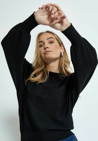 Schwarzer gestrickter Pullover mit langen, ballonförmigen Ärmeln. Angenehme Textur, runder Ausschnitt und gerippte Bündchen. Das Modell posiert mit erhobenen Armen und zeigt die Passform.