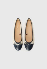 Paire de ballerines crème et noires avec petits nœuds à l'avant et semelles beiges sur fond blanc.
