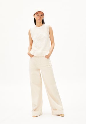 Gebreide mouwloze top in off-white, met een geribbelde zoom. Gecombineerd met losse beige broeken, iets opgerold bij de enkels, en eenvoudige schoenen.