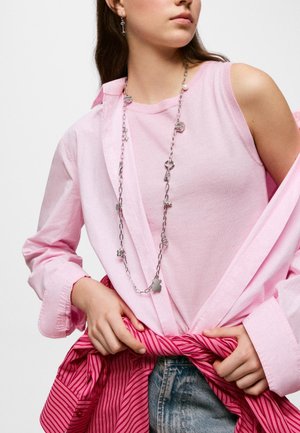 Giovane donna che indossa un top senza maniche rosa chiaro sovrapposto a una camicia rosa a maniche lunghe, una collana con charm in argento e tiene una giacca rosa a righe in vita.
