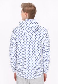 Giacca a vento con cappuccio in tessuto bianco con un motivo astratto blu ripetuto. Presenta polsini elasticizzati e una vestibilità rilassata.