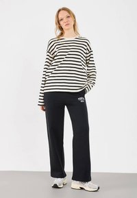 Γυναίκα ντυμένη με μαύρο παντελόνι με φαρδιές γάμπες που έχει το κείμενο "Tokyo 1963", ένα κρεμ και μαύρο ριγέ πουκάμισο με μακριά μανίκια και λευκά αθλητικά παπούτσια, ενώ στέκεται σε λευκό φόντο.