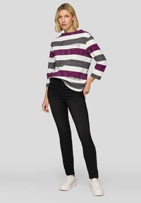 Gestreiftes Oberteil in Weiß und Grau mit lila Akzenten, 3/4-Ärmeln und grafischen Details. Kombiniert mit schwarzen Jeans und weißen Sneakers.