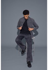 Grå denimjacka över en svart t-shirt, i kombination med mörkgrå vida jeans och svarta skor, sittande på en pall mot en grå bakgrund.