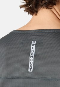 Dunkelgraues Sportshirt mit einem Gittermuster. Es hat ein vertikales weißes Etikett mit der Aufschrift "SWEDEMOUNT" am hinteren Nackenbereich.