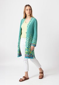 Turkiksin pitkä neuletakki, jossa on geometrisia kuvioita ja monivärisiä yksityiskohtia hihoissa. Yhdistetty vaaleankeltaisen topin ja valkoisten housujen kanssa.