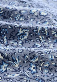 Ziedu audums ar zilu un bēšu ziedu rakstiem uz gaiši pelēka fona, ar teksturētu, austas virsmas un maigu nolikumu.