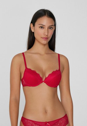 Soutien-gorge en dentelle rouge avec un motif en relief, support à armatures et détails décoratifs aux bretelles et au centre avant. Fermeture au dos.