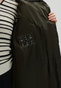 Olijfgroene pufferjack met een gladde, glanzende textuur. De binnenlabels geven de kenmerken aan: ademend, waterafstotend en lichtgewicht.